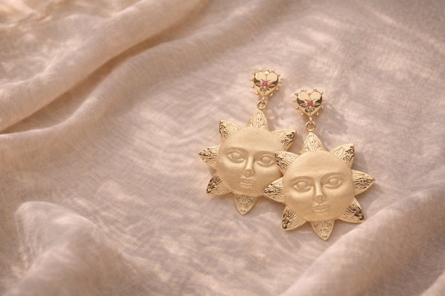 Jane Win STRONG Under the Sun gold drop earrings with sun face design and pink tourmaline heart posts styled on soft linen fabric